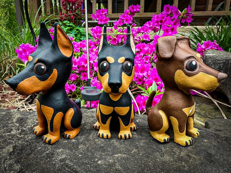 May include: Three small, hand-painted dog figurines. The dogs are black, brown, and tan with white accents. They are sitting on a gray, textured surface.