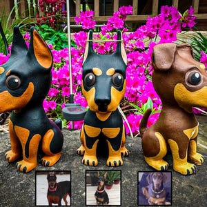 May include: Three hand-painted ceramic dog figurines, each with a different color scheme. The figurines are black and tan, brown and tan, and black and white. The figurines are sitting on a rock surface with pink flowers in the background.
