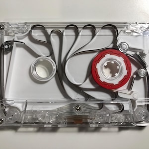 May include: A clear plastic cassette tape with a white interior frame and a single red spool. The black magnetic tape is visible, looping through the internal mechanism. The device features four small metal screws at the corners and centre.