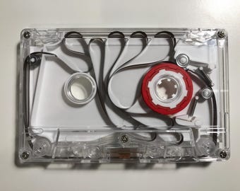May include: A clear plastic cassette tape with a white interior frame and a single red spool. The black magnetic tape is visible, looping through the internal mechanism. The device features four small metal screws at the corners and center.