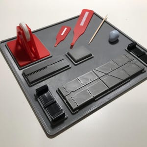 Op de afbeelding: Een grijs vierkant organisator-dienblad met diverse uitsparingen waarin een rode tape-dispenser, twee rode plastic gereedschappen, een houten tandenstoker, een kleine lichtblauwe kleibal en diverse zwarte en grijze modulaire kunststof onderdelen liggen.