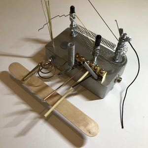 May include: A silver metal box with various wires, springs, and other metal objects attached to it. A wooden stick is attached to the box.