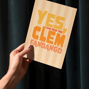 May include: A rectangular greeting card with a striped background in shades of beige and orange. The card features the words "YES, I CAN HEAR YOU CLEM FANDANGO" in bold, orange and yellow lettering. The card is held by a person.