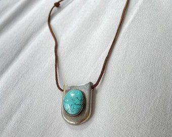 May include: A silver pendant necklace with a turquoise stone set in a silver frame. The pendant hangs from a brown cord necklace with a silver clasp.