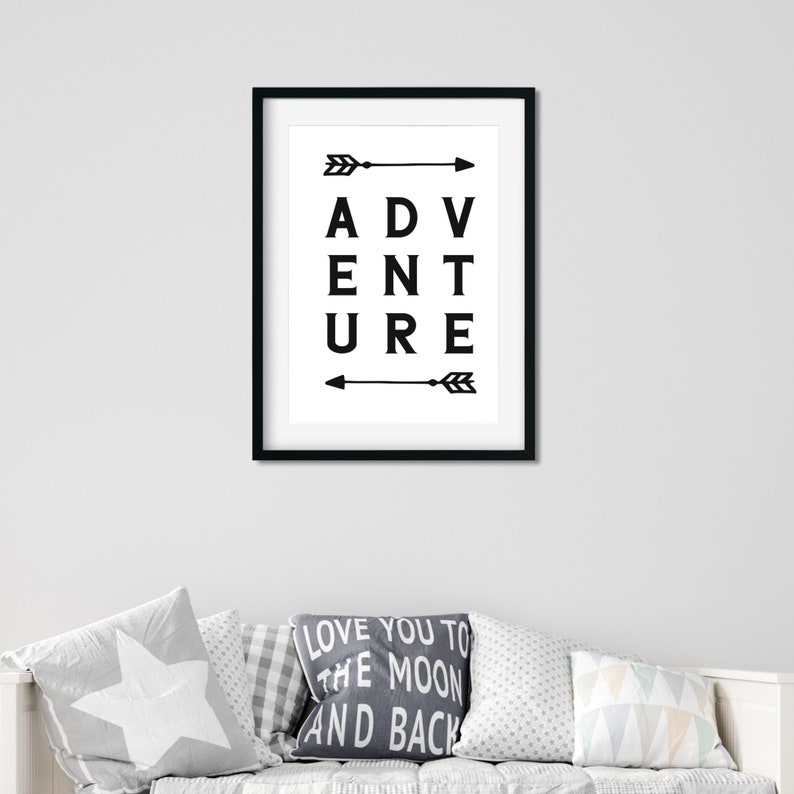 May include: Black framed wall art with the word "ADVENTURE" in bold, black capital letters. Arrows point left and right. The artwork is displayed above a white couch with decorative pillows.