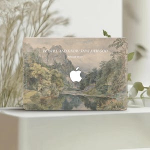 Puede incluir: Funda para portátil con una pintura de paisaje que representa un río, árboles y montañas. El logotipo de Apple es visible. El texto dice "BE STILL AND KNOW THAT I AM GOD" y "PSALM 46:10". La obra de arte utiliza colores apagados.