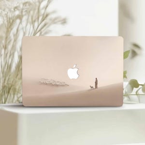 Può includere: Una custodia beige per laptop con una serena scena desertica che raffigura una persona, un cane e un gregge di pecore. Il logo Apple è ben visibile. Il design evoca un senso di tranquillità e avventura.