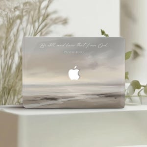 Puede incluir: Una funda para portátil con una pintura de paisaje en tonos beige, gris y blanco. El diseño incluye el logotipo de Apple y el texto "Be still, and know that I am God. Salmo 46:10."