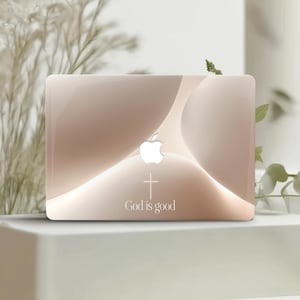 以下が含まれることがあります： ベージュと白のグラデーションデザインのノートパソコンケース。Appleのロゴ、白い十字架、そして「God is good」の文字が見えます。ケースは光沢のある仕上がりです。