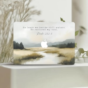 Puede incluir: Una funda para portátil con una pintura de paisaje de un río, árboles y un cielo nublado. La funda tiene el logotipo de Apple y el texto: «Me guía junto a aguas tranquilas. Restaura mi alma. Salmo 23:2-3."