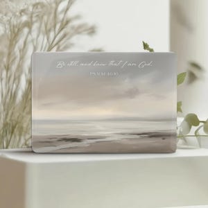May include: A rectangular plaque featuring a soft, muted landscape painting of a beach at dusk with beige, gray, and white tones. The text "Be still, and know that I am God. PSALM 46:10" is printed in a white, elegant script font across the upper portion.