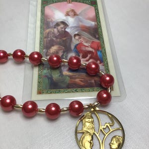 May include: A red beaded rosary with gold-colored accents and a gold pendant featuring religious figures. A religious card with an image of biblical figures is behind the rosary.
