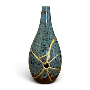 May include: A teardrop-shaped ceramic vase featuring a mottled blue and brown glaze. The surface is accented with prominent gold-colored lines that radiate from a small circular opening, creating a distinct pattern across the body of the vessel.