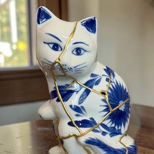 May include: White ceramic cat figurine with blue floral patterns and gold-colored kintsugi-style repairs. The cat has blue accents on its ears and eyes. The figurine is sitting on a wooden surface.