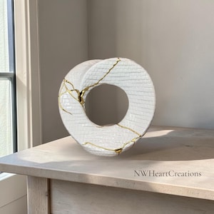 May include: White ceramic sculpture with a circular design, featuring gold accents along the cracks. The sculpture has a textured surface and is placed on a light-coloured wooden surface. The text "NWHeartCreations" is visible.