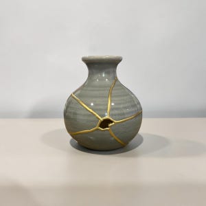 May include: A small, round, grey ceramic vase with a narrow neck and a wide body. The surface features a series of thin, gold-colored lines radiating from a central point, creating a repair-like pattern across the smooth, glossy finish.