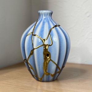 Puede incluir: Un jarrón de cerámica a rayas azules y blancas con líneas de reparación kintsugi doradas. El jarrón tiene forma redondeada y cuello estrecho. Las líneas doradas resaltan las grietas.