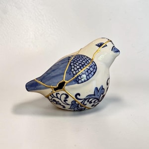 May include: A ceramic bird figurine featuring a white base with blue painted patterns on the wings and body. The surface displays gold-colored lines tracing cracks across the form, highlighting a repair technique that connects the segments.