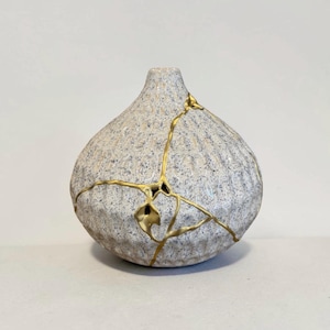 May include: A small, round ceramic vase with a textured, speckled light gray and white surface. The vase features prominent gold-colored lines that trace across its body, highlighting a series of cracks in the ceramic material.