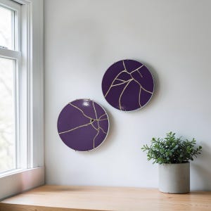 May include: Two round, purple wall art pieces with gold-colored crack patterns. The art has a silver metal frame and is mounted on a white wall. A small potted plant sits on a wooden surface below the art.
