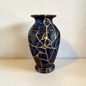 May include: A deep blue ceramic vase featuring a network of gold-colored lines across its surface. The vase has a flared rim, a rounded body, and a pedestal base. The gold lines create a branching pattern that resembles cracks across the dark blue.