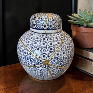 Peut inclure: Un pot en céramique rond bleu et blanc avec un couvercle, décoré d'un motif floral. Le pot présente des lignes dorées, clin d'œil à l'art japonais du Kintsugi. Le pot est posé sur une surface en bois.