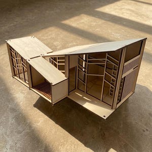 May include: A small architectural model made from light-coloured wood. The model has several sections, including a sloping roof, window frames, and open spaces, demonstrating a modern design. The model is placed on a neutral surface.