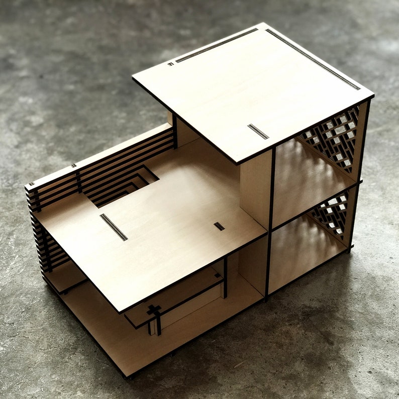 May include: A wooden model of a house with a flat roof and a lattice-like design on the side. The model is made of light brown wood and has black lines outlining the different sections.