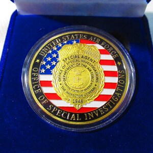 Może przedstawiać: Złoty medal z amerykańską flagą w kolorze niebieskim, białym i czerwonym. Medal zawiera napis "United States Air Force Office of Special Investigations" i "Special Agent" na krawędzi. W centrum medalu znajduje się pieczęć z napisem "Department of the Air Force" i "The Inspector General 1948".