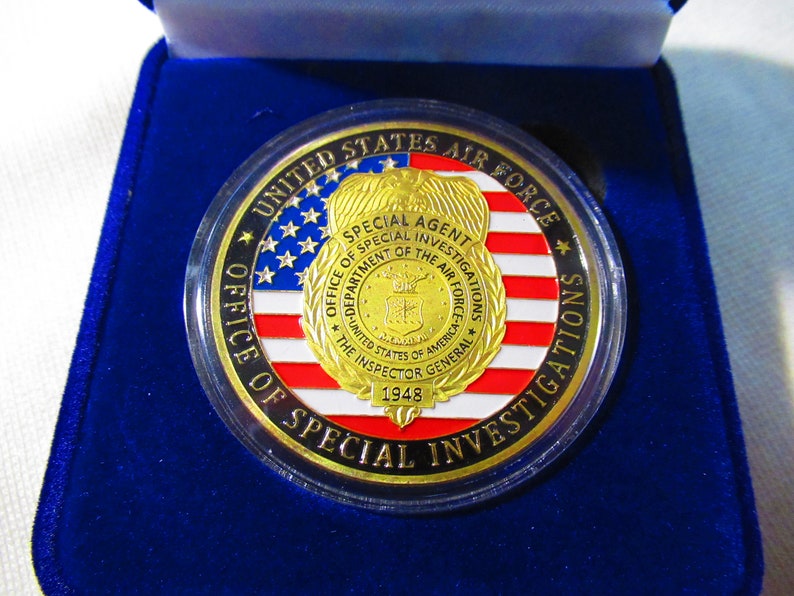 Może przedstawiać: Złoty medal z amerykańską flagą w kolorze niebieskim, białym i czerwonym. Medal zawiera napis "United States Air Force Office of Special Investigations" i "Special Agent" na krawędzi. W centrum medalu znajduje się pieczęć z napisem "Department of the Air Force" i "The Inspector General 1948".
