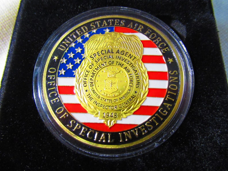 Może przedstawiać: Złota moneta z wzorem amerykańskiej flagi w kolorze czerwonym, białym i niebieskim. Na monecie widnieje tekst "United States Air Force Office of Special Investigations Special Agent" oraz "The Inspector General 1948".
