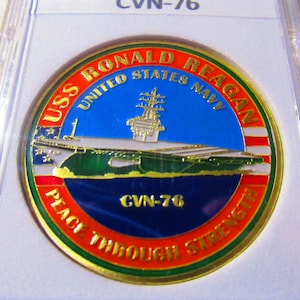 Op de afbeelding: Een gouden challenge-munt met een rood, wit en blauw Amerikaans vlagontwerp. De munt toont een afbeelding van de USS Ronald Reagan vliegdekschip met de tekst "USS Ronald Reagan United States Navy" en "CVN-76 Peace Through Strength".