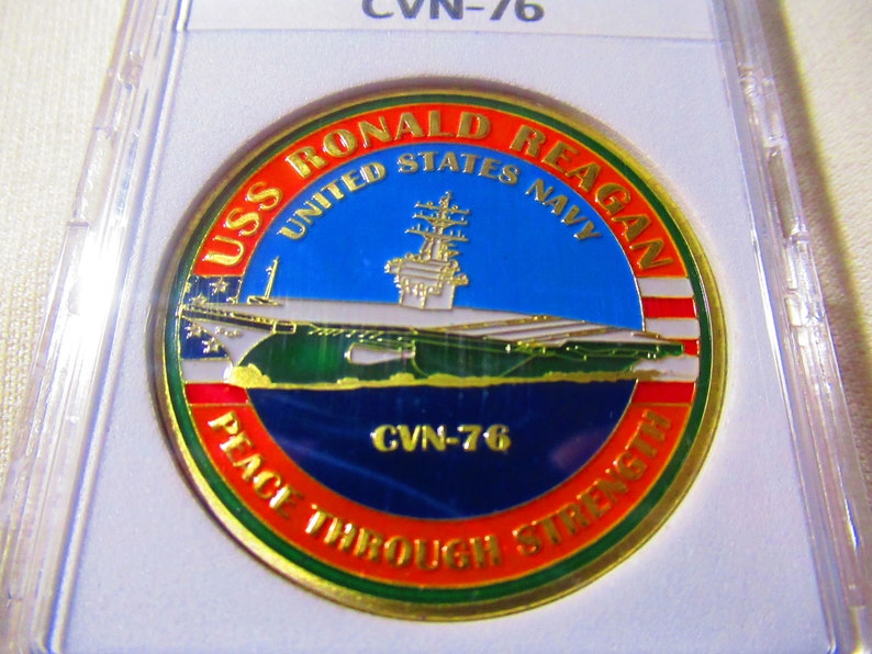 Op de afbeelding: Een gouden challenge-munt met een rood, wit en blauw Amerikaans vlagontwerp. De munt toont een afbeelding van de USS Ronald Reagan vliegdekschip met de tekst "USS Ronald Reagan United States Navy" en "CVN-76 Peace Through Strength".
