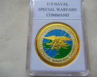 May include: A gold-colored coin with a white, blue, and green design. The coin features the words "United States Naval Special Warfare Command" and a stylized image of a seabird with a trident.