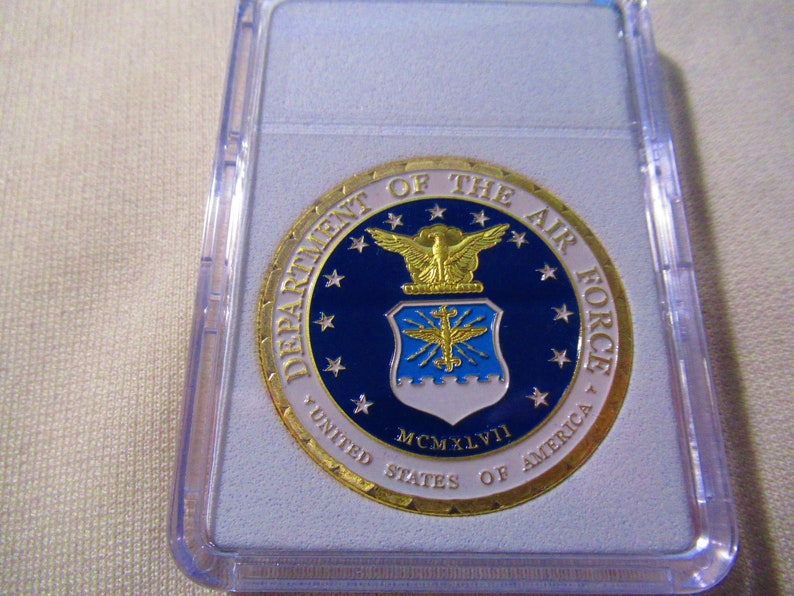 Może przedstawiać: Złota moneta z niebiesko-białym godłem Sił Powietrznych Stan&oacute;w Zjednoczonych. Na monecie widnieją napisy "Department of the Air Force" i "United States of America". Na monecie widnieje r&oacute;wnież rok "MCMXLVI".