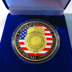 Może przedstawiać: Złoty medal z czerwono-biało-niebieskim wzorem amerykańskiej flagi. Medal zawiera napis "UNITED STATES AIR FORCE" i "OFFICE OF SPECIAL INVESTIGATIONS". W centrum medalu znajduje się złota odznaka z napisem "SPECIAL AGENT" i "DEPARTMENT OF THE AIR FORCE".