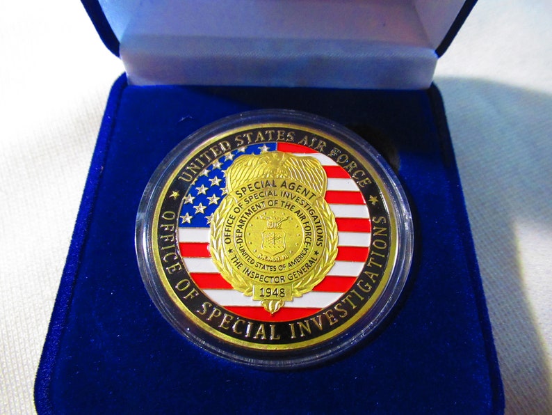 Może przedstawiać: Złoty medal z czerwono-biało-niebieskim wzorem amerykańskiej flagi. Medal zawiera napis "UNITED STATES AIR FORCE" i "OFFICE OF SPECIAL INVESTIGATIONS". W centrum medalu znajduje się złota odznaka z napisem "SPECIAL AGENT" i "DEPARTMENT OF THE AIR FORCE".