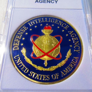 Może przedstawiać: Złota moneta z niebieskim tłem i napisem "Defense Intelligence Agency United States of America" wokół krawędzi. W centrum monety znajduje się złoty glob z czerwoną wstążką wokół niego i pochodnią nad nim.