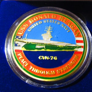 Op de afbeelding: Een gouden munt met een blauwe, witte en groene afbeelding van de vliegdekschip USS Ronald Reagan. De munt heeft de tekst "USS Ronald Reagan", "United States Navy", "CVN-76" en "Peace Through Strength" rond de rand.