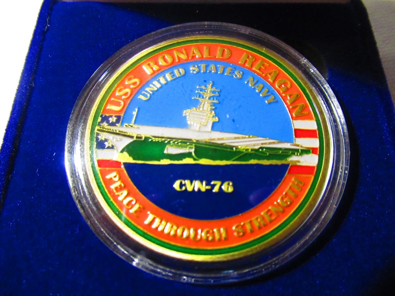 Op de afbeelding: Een gouden munt met een blauwe, witte en groene afbeelding van de vliegdekschip USS Ronald Reagan. De munt heeft de tekst "USS Ronald Reagan", "United States Navy", "CVN-76" en "Peace Through Strength" rond de rand.