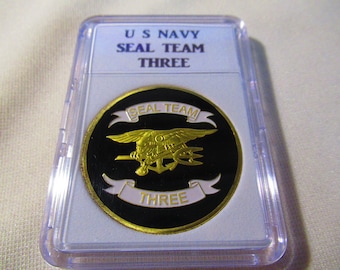 Pode incluir: Uma moeda dourada com fundo preto que apresenta um emblema de equipe SEAL em dourado com as palavras "SEAL TEAM THREE" em letras brancas.