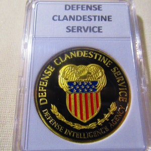 Puede incluir: Una moneda de color dorado con fondo negro y las palabras "Defense Clandestine Service Defense Intelligence Agency" alrededor del borde. El centro de la moneda presenta un águila dorada con un escudo con rayas rojas y blancas y estrellas azules.