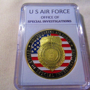 Może przedstawiać: Złoty medal z czerwono-biało-niebieskim wzorem amerykańskiej flagi. Medal zawiera napis "United States Air Force" i "Office of Special Investigations" wok&oacute;ł krawędzi. W centrum medalu znajduje się odznaka z napisem "Special Agent" i "Office of Special Investigations, Department of the Air Force".