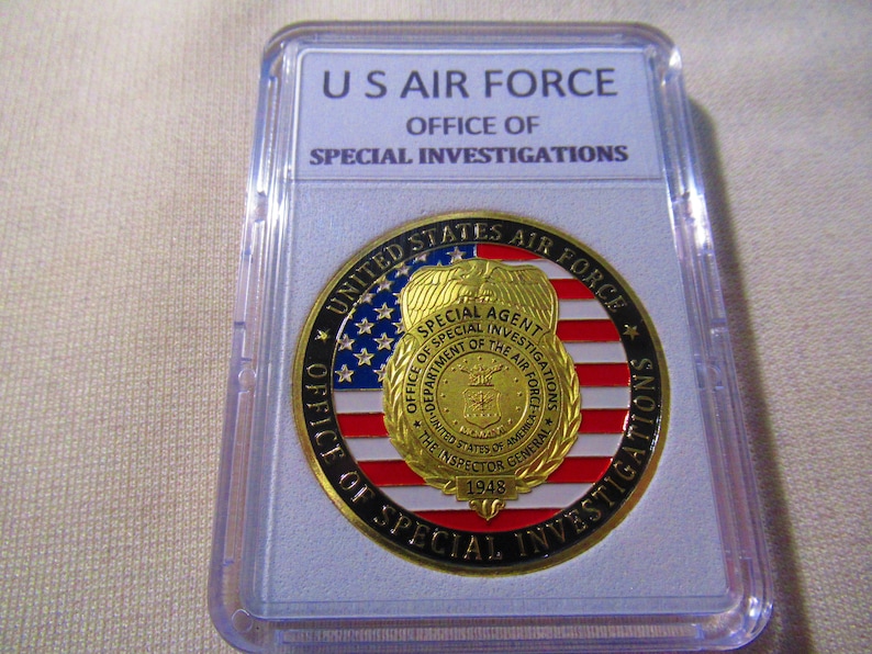 Może przedstawiać: Złoty medal z czerwono-biało-niebieskim wzorem amerykańskiej flagi. Medal zawiera napis "United States Air Force" i "Office of Special Investigations" wok&oacute;ł krawędzi. W centrum medalu znajduje się odznaka z napisem "Special Agent" i "Office of Special Investigations, Department of the Air Force".