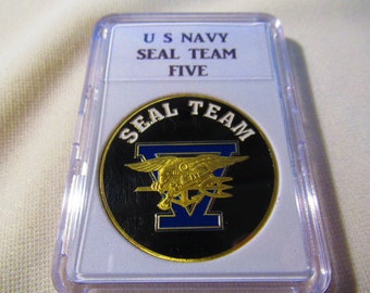 May include: A gold coin with a black background and a gold seal team emblem. The emblem features a stylized eagle with a trident and the number five in blue. The coin is in a clear plastic case with the text "US Navy Seal Team Five" on the label.