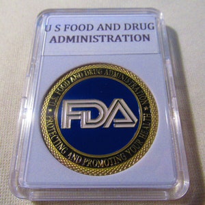 May include: A gold-colored coin with a blue and white FDA logo. The coin is surrounded by a gold-colored rope design and the text "U.S. Food and Drug Administration Protecting and Promoting Your Health".