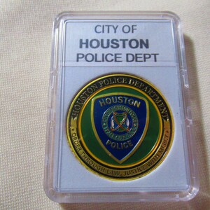 May include: A gold-colored coin with a green and blue shield design featuring the words "Houston Police Department" and "Houston Police" in a white circle. The coin is in a clear plastic case with a white background that reads "City of Houston Police Dept."