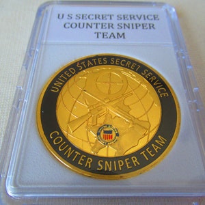 Puede incluir: Una moneda de color dorado con un borde negro y el texto "United States Secret Service Counter Sniper Team" alrededor del borde. El centro de la moneda presenta un globo terráqueo con rifles cruzados y una mira.