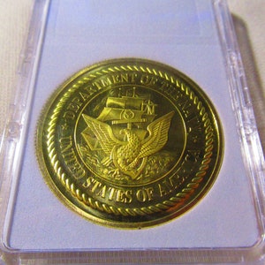 Op de afbeelding: Een gouden munt met de inscriptie "Department of the Navy United States of America" en een adelaar met een anker in zijn klauwen.