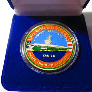 Op de afbeelding: Een gouden munt met een blauw, groen en rood ontwerp. De munt toont een afbeelding van de vliegdekschip USS Ronald Reagan en de tekst "USS Ronald Reagan United States Navy CVN-76 Peace Through Strength".